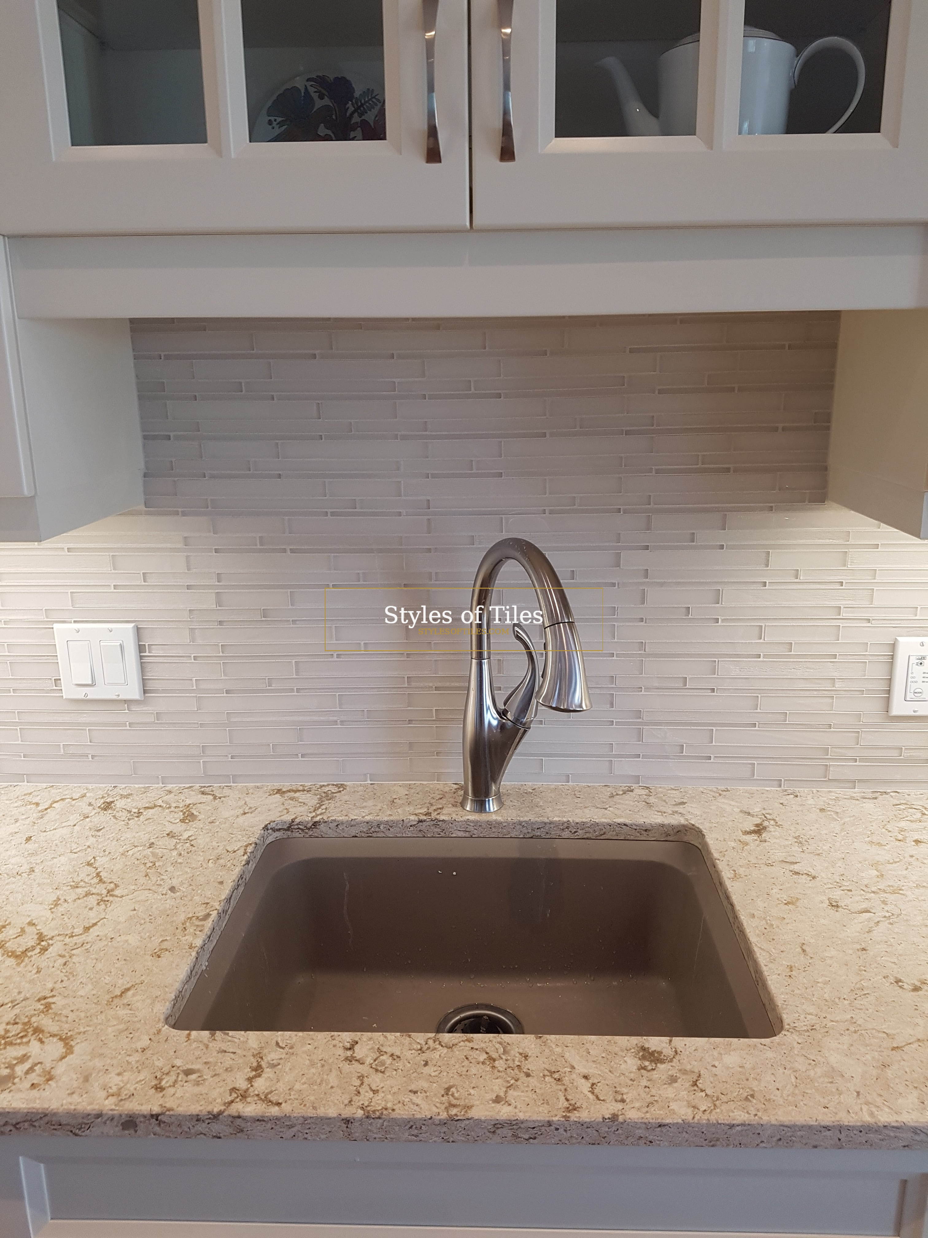 White stone strip-mosaic backsplash behind a kitchen sink, tiled up under glass-door cabinets