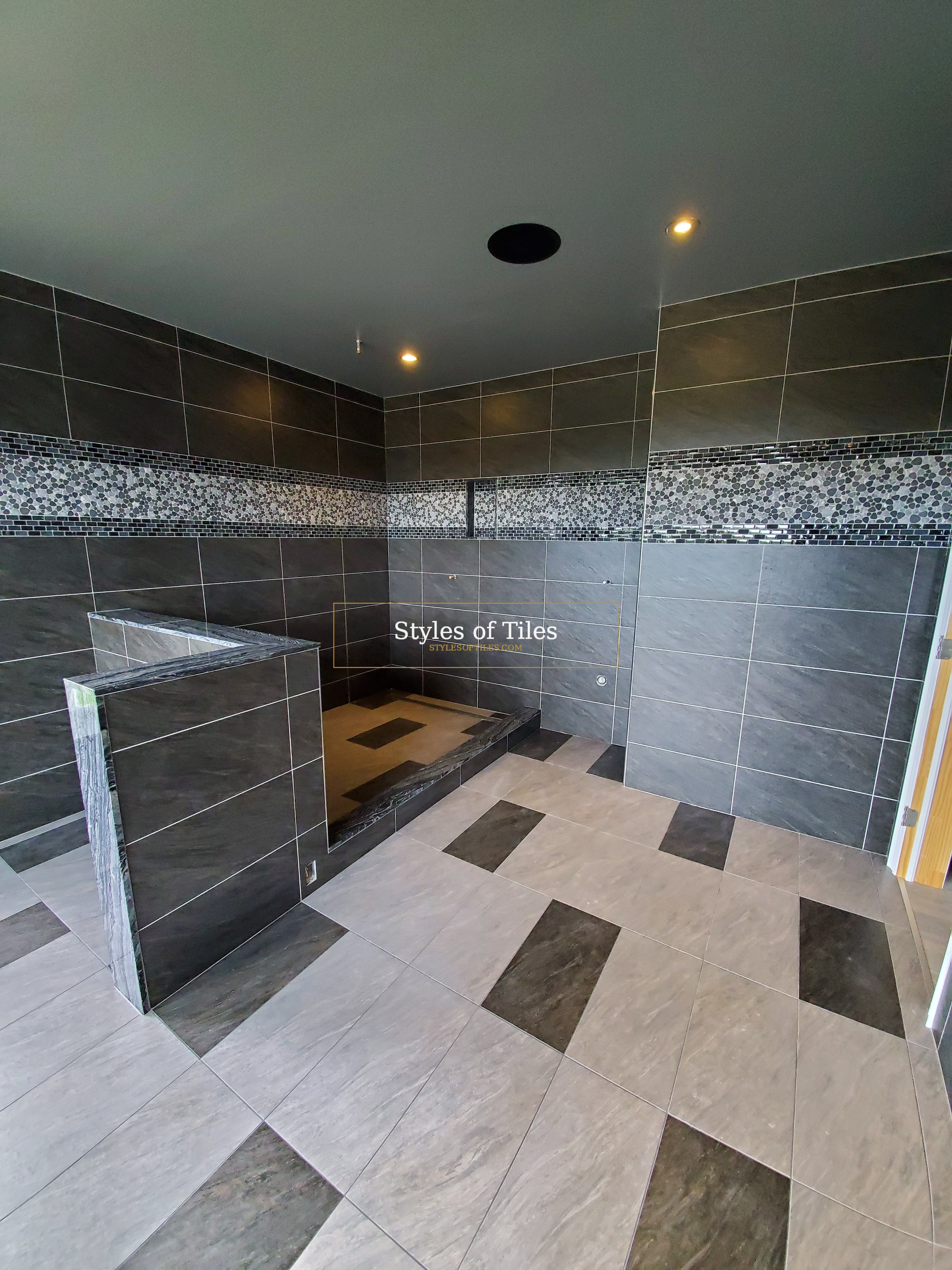 Large open bathroom with light grey floor tile, dark accent tiles and a tiled half-wall at the shower